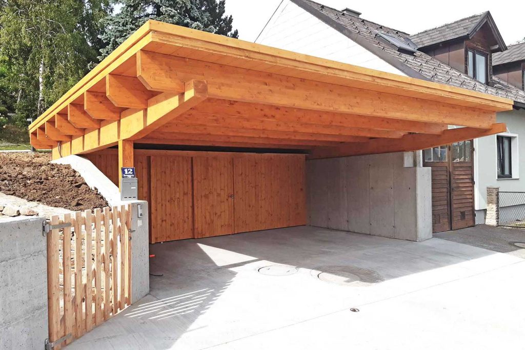 Carport aus Holz mit weit auskragendem Dach und Holzwand neben einem Wohnhaus, davor ein Holzzaun
