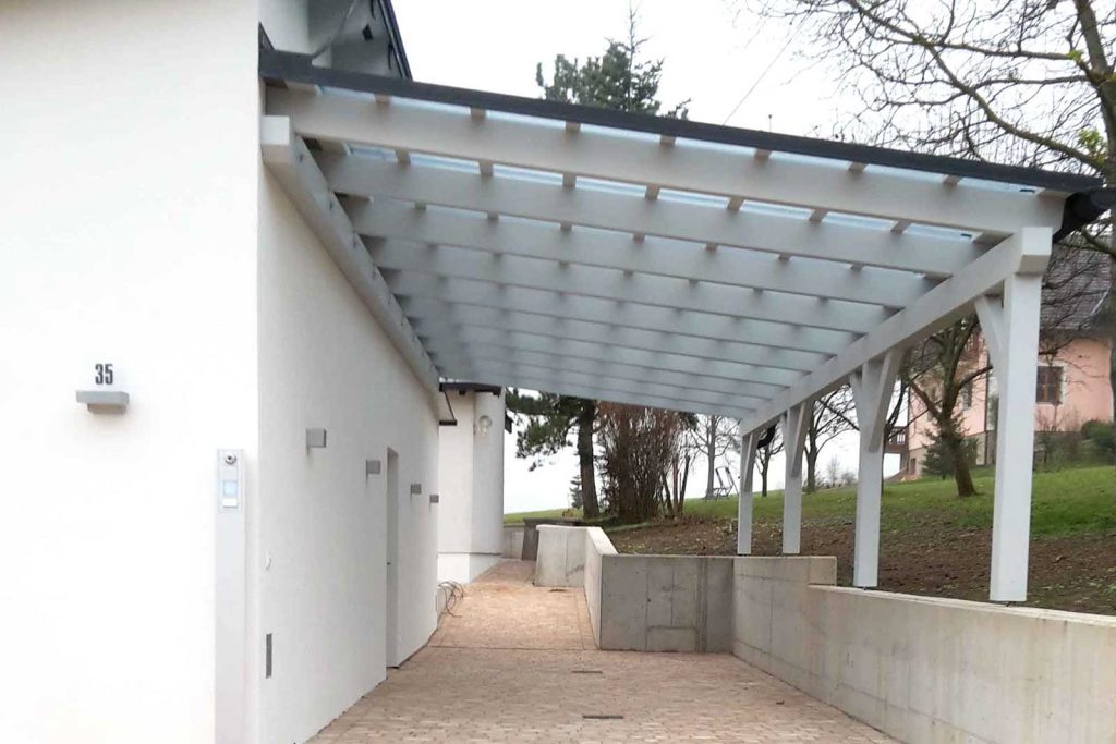 Carport aus Holz mit hellem, lichtdurchlässigem Dach auf weiß gestrichenen Stützen entlang einer Hauswand
