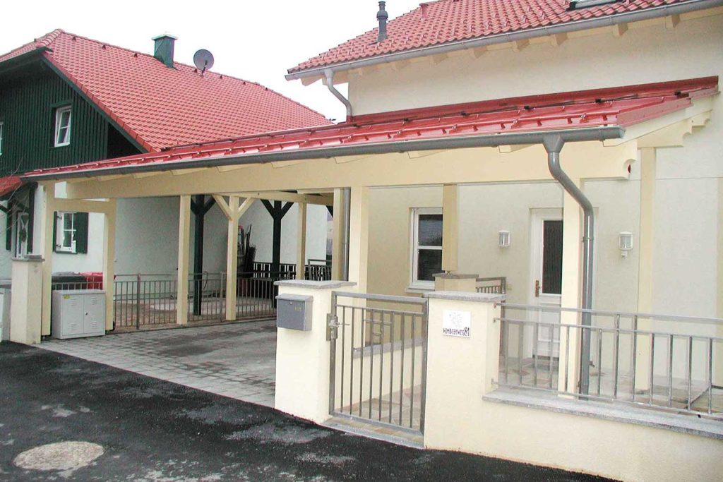 Carport aus Holz mit rotem Dach als Anbau an ein Wohnhaus, daneben Einfahrt und Gartenzaun