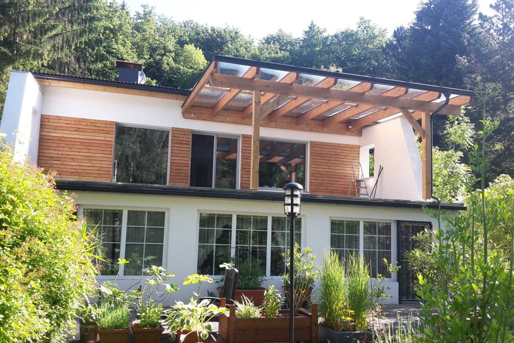 Pergola aus Holz über einer Terrasse an einem Wohnhaus mit Holzverkleidung, davor ein bepflanzter Garten