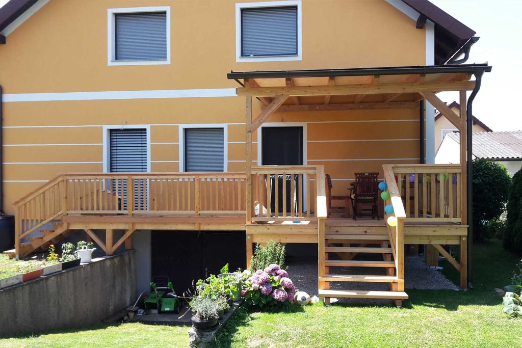 Holzterrasse mit Treppe und Geländer unter einer Terrassenüberdachung aus Holz an einem gelben Wohnhaus