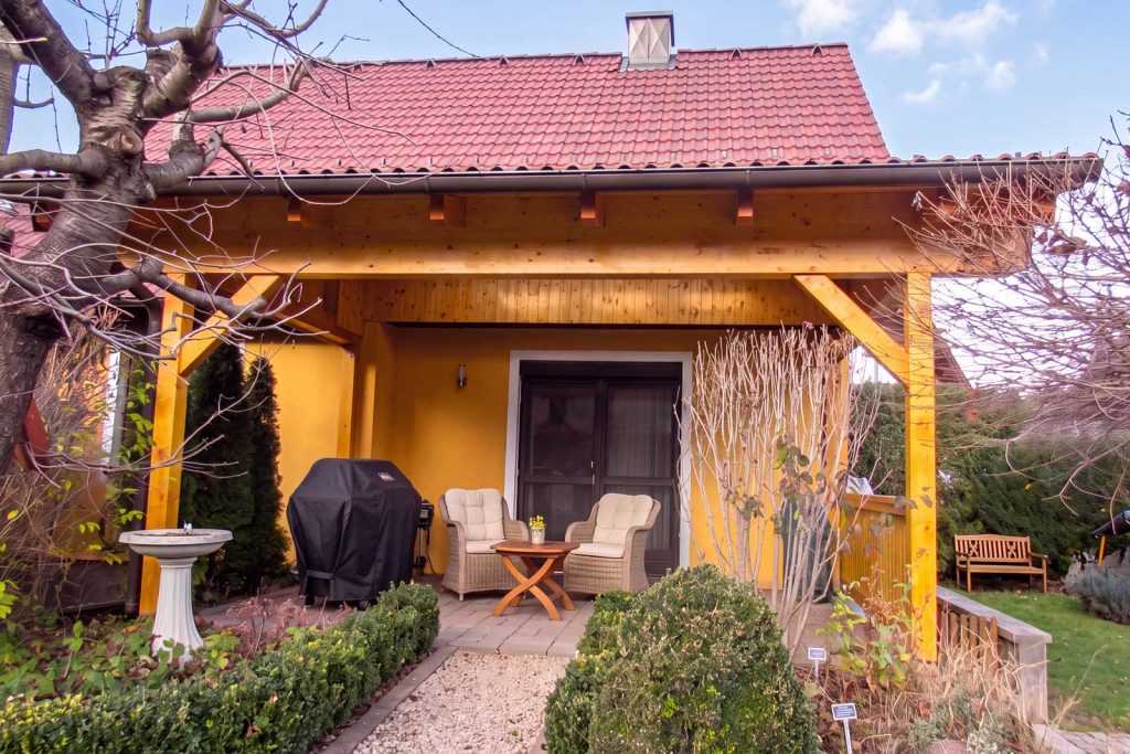 Terrassenüberdachung aus Holz an einem Wohnhaus mit rotem Ziegeldach, darunter ein Sitzplatz mit Gartenmöbeln